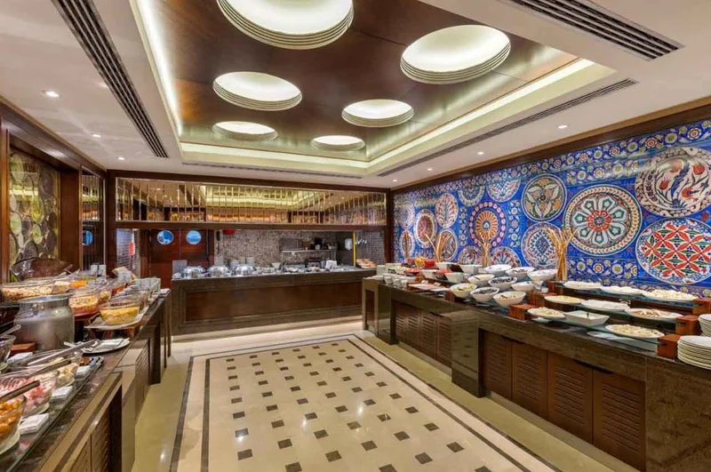 سالن صبحانه سلف‌سرویس هتل تایتانیک سیتی تکسیم، با میزهای بوفه متنوع و دیواری که با کاشی‌کاری‌های سنتی و رنگارنگ ترکی تزئین شده است.